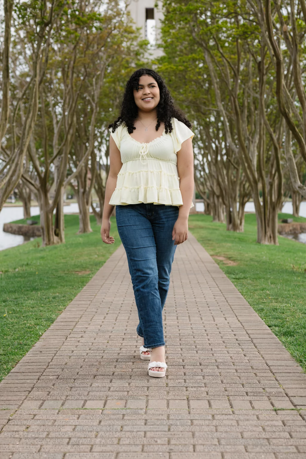 Nolan's senior photos taken at Furman University: A young woman with curly hair wearing a light yellow blouse and blue jeans walks along a paved path lined with trees in a park.