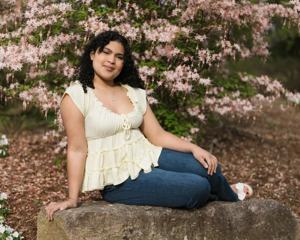 Nolan's senior photos taken at Furman University: A person with curly hair, wearing a light yellow top and blue jeans, sits on a large rock in front of blooming pink flowers in an outdoor garden.