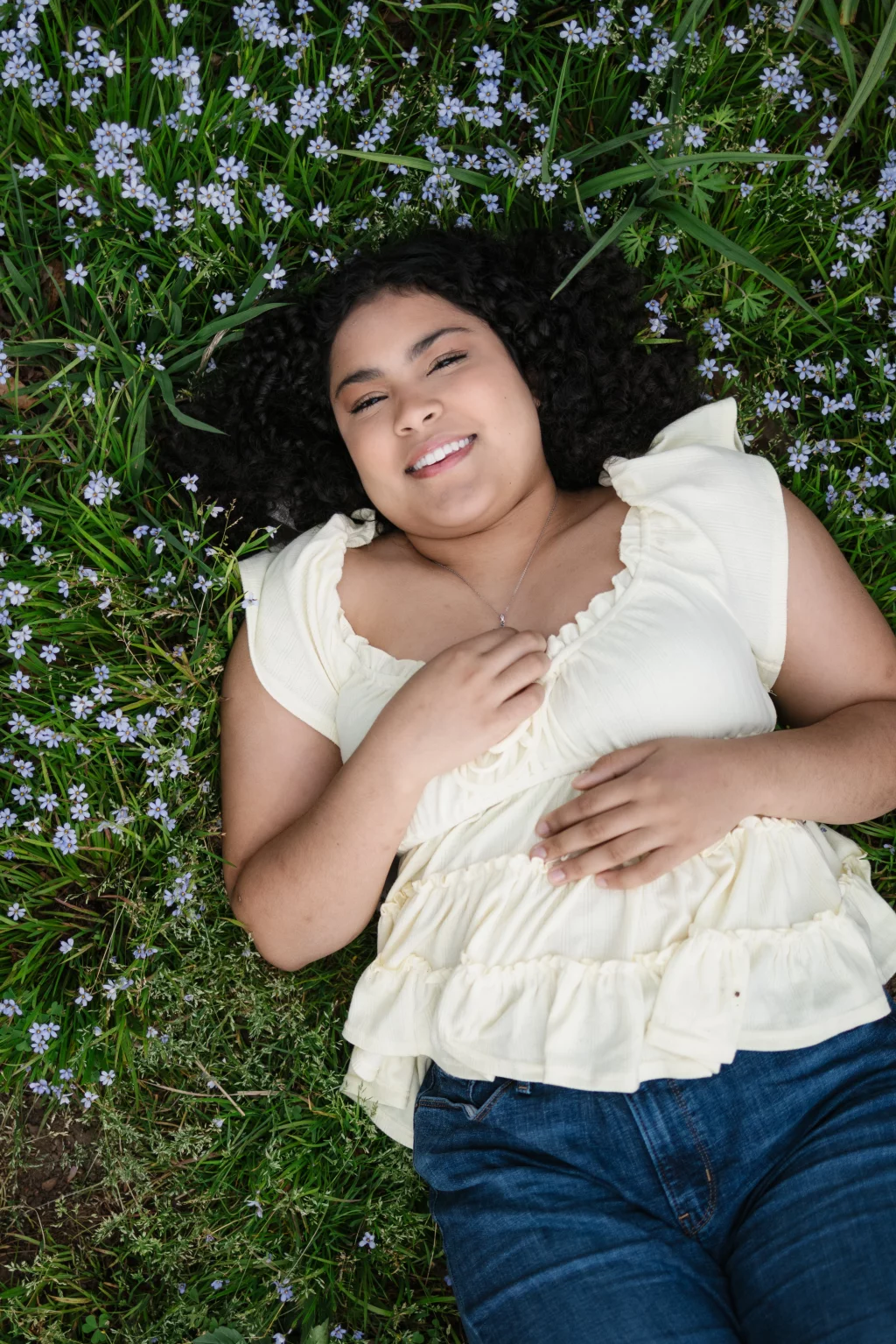 Nolan's senior photos taken at Furman University: Person with curly hair wearing a cream ruffled blouse and jeans lying on grass with small purple flowers, smiling at the camera.
