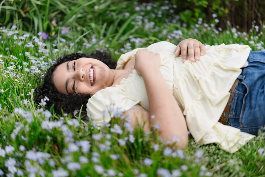 Nolan's senior photos taken at Furman University: A smiling person lies on their back in a field of green grass and small purple flowers, wearing a light yellow top and blue jeans.