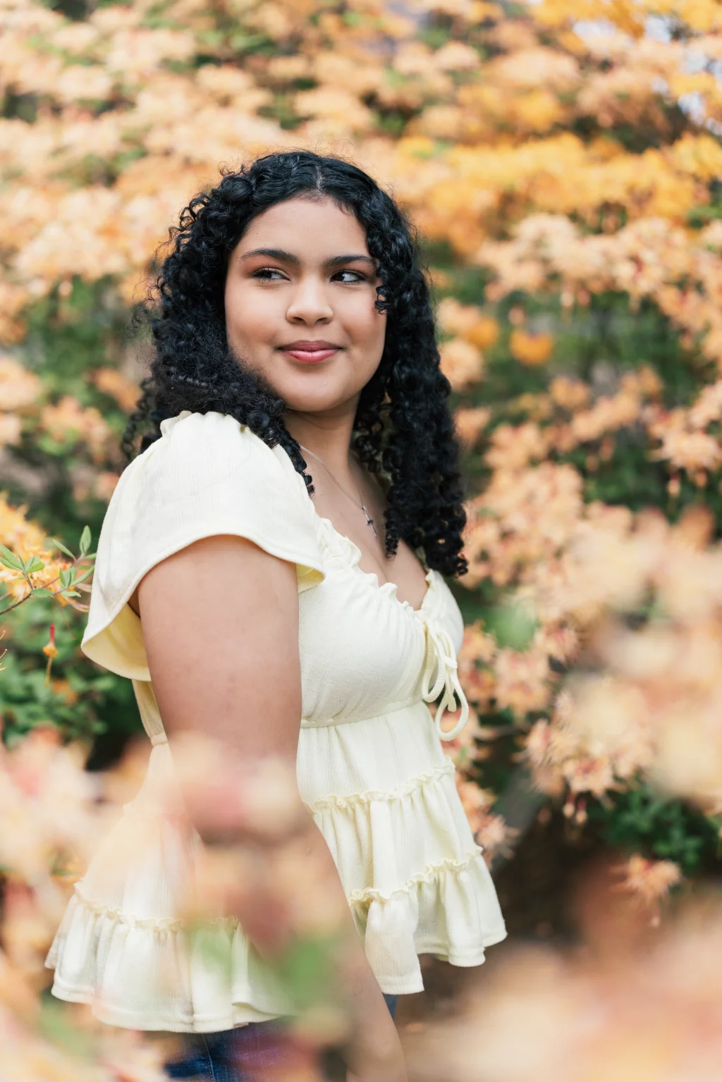 Nolan's senior photos taken at Furman University: Woman with curly hair wearing a pale yellow top standing outdoors, surrounded by soft-focus yellow and orange flowering bushes.