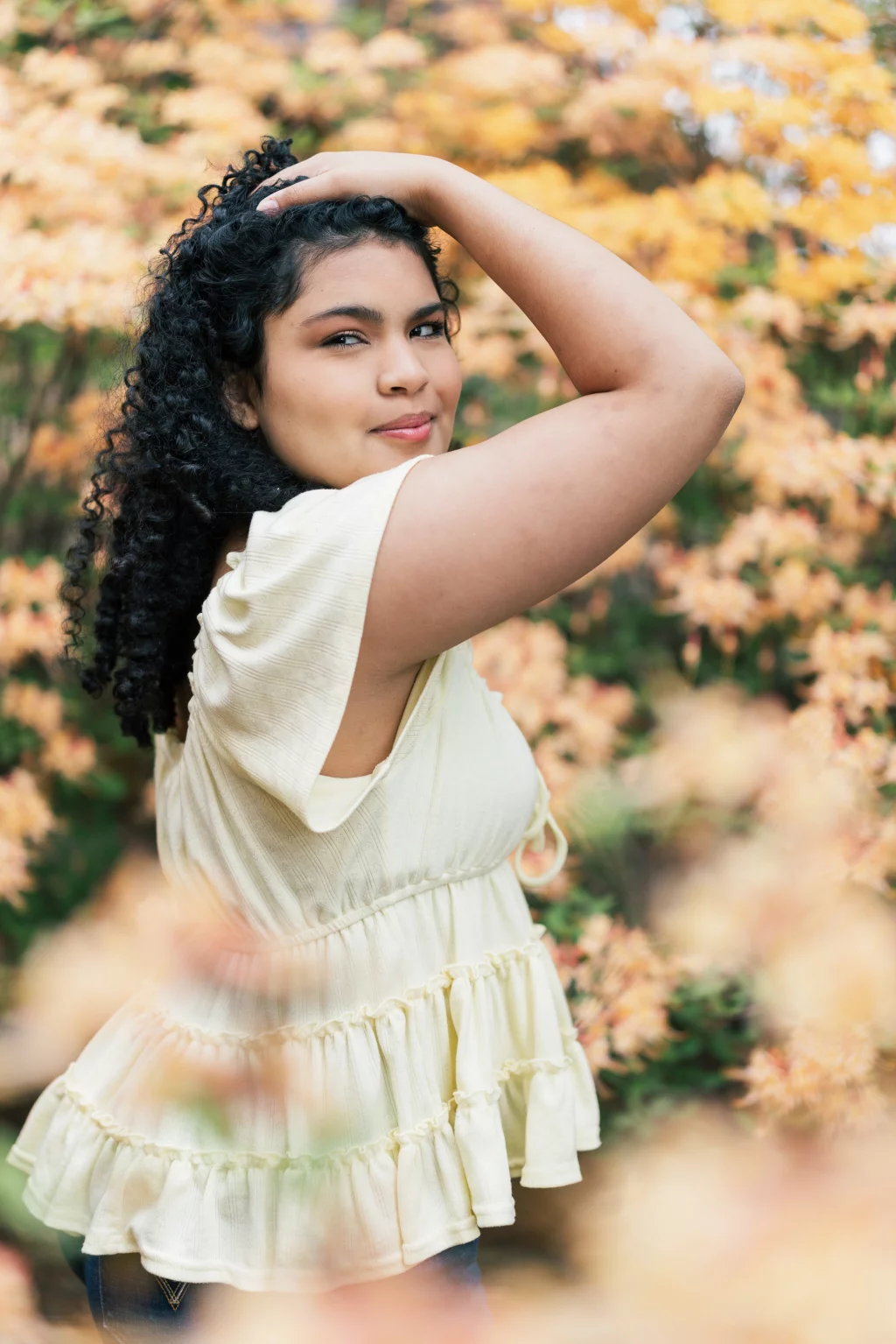 Nolan's senior photos taken at Furman University: Woman with curly hair wearing a light yellow ruffled top poses outdoors with one arm raised over her head, standing in front of blurred yellow and green foliage.