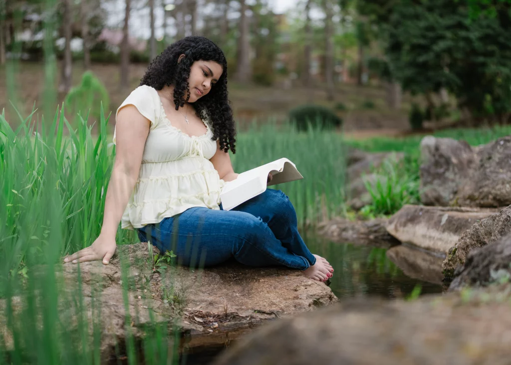 Nolan's senior photos taken at Furman University: A woman with curly hair sits barefoot on a rock beside a stream, reading a book in a lush, green outdoor setting.