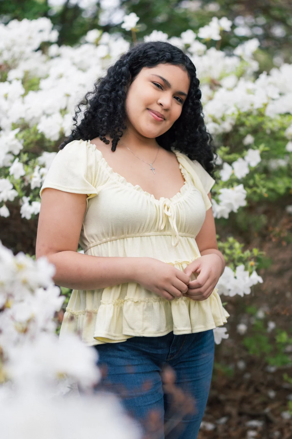 Nolan's senior photos taken at Furman University: Smiling young woman with curly hair standing among blooming white flowers, wearing a light yellow top and blue jeans.