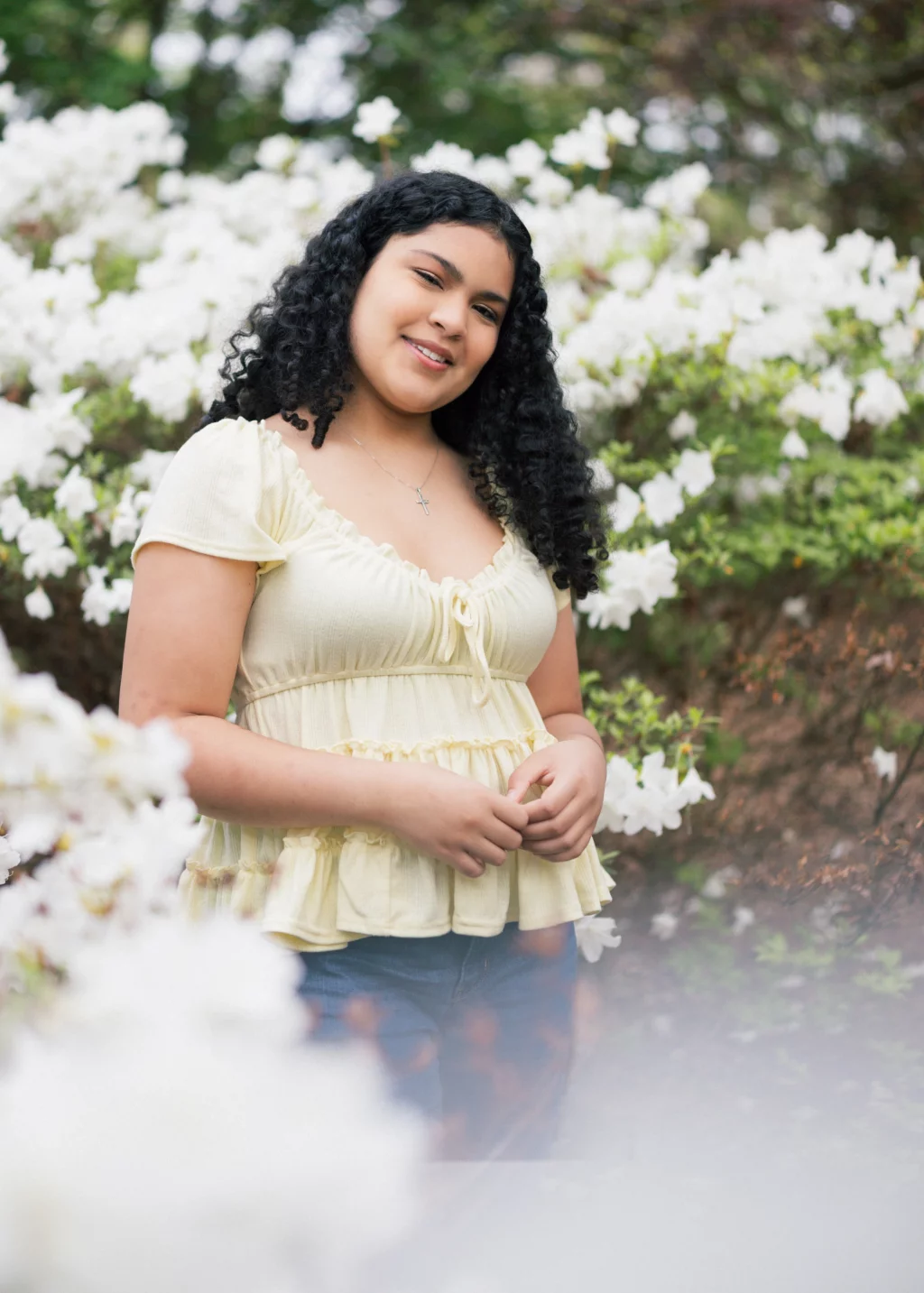 Nolan's senior photos taken at Furman University: Young woman with curly hair wearing a light yellow blouse, standing outdoors in front of blooming white flowers and greenery, smiling at the camera.