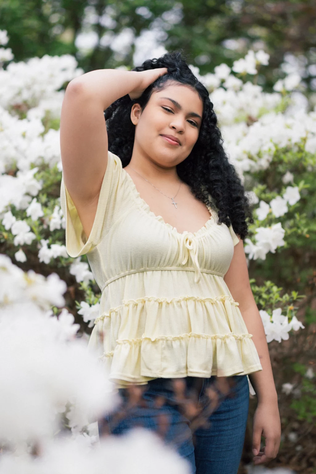 Nolan's senior photos taken at Furman University: A young woman with curly hair stands outdoors in front of blooming white flowers, wearing a light yellow ruffled blouse and blue jeans, with one hand resting on her head and a slight smile on her face.