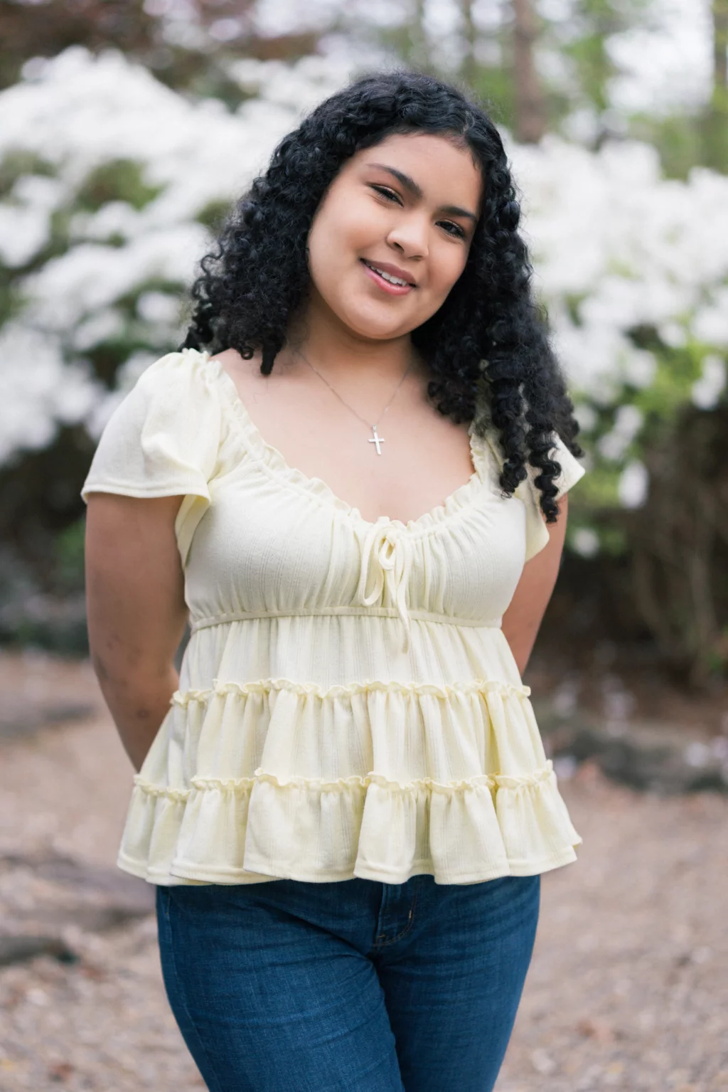 Nolan's senior photos taken at Furman University: Young woman with curly black hair wearing a light yellow ruffled blouse and blue jeans, standing outdoors in front of blooming white flowers, smiling at the camera.