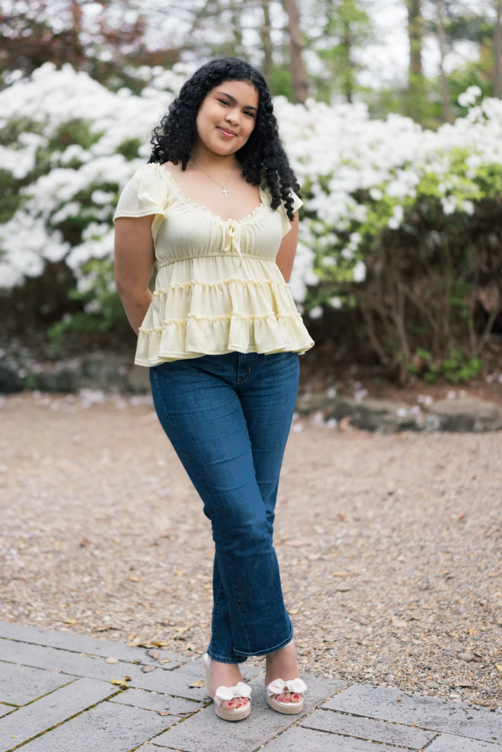 Nolan's senior photos taken at Furman University: A young woman with curly hair stands outdoors in front of white flowering bushes. She is wearing a light yellow ruffled top, blue jeans, and cream-colored wedge sandals with bows. She is smiling slightly with her hands behind her back.