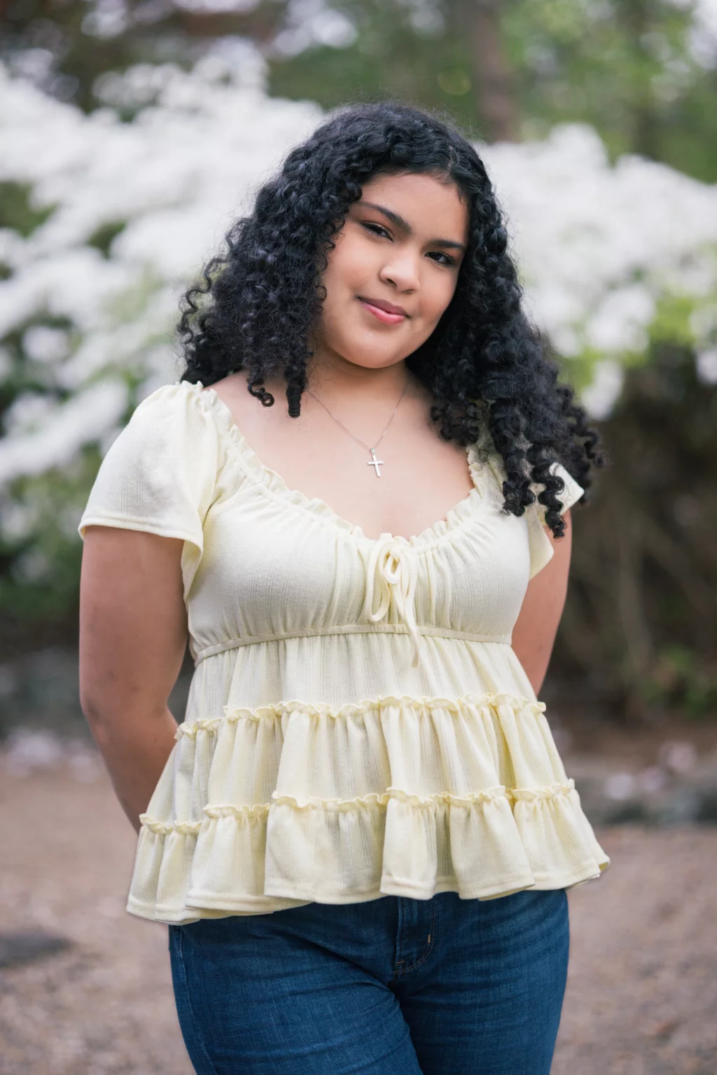 Nolan's senior photos taken at Furman University: A young woman with curly hair stands outdoors, wearing a light yellow ruffled top, blue jeans, and a silver cross necklace, with white flowers and greenery in the background.