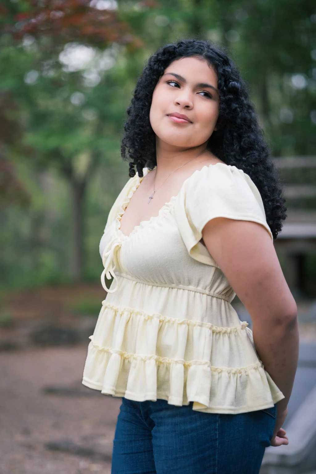 Nolan's senior photos taken at Furman University: Young woman with curly hair wearing a light yellow ruffled blouse and blue jeans, standing outdoors with green trees in the background.