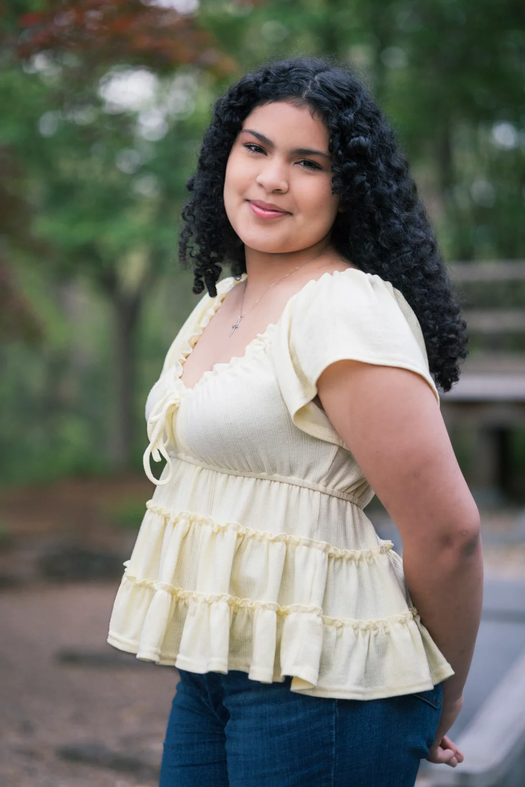 Nolan's senior photos taken at Furman University: Young woman with curly dark hair wearing a pale yellow ruffled blouse and blue jeans, standing outdoors and smiling at the camera with a green, blurred background.