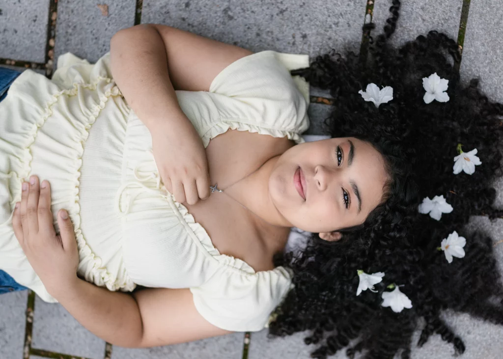 Nolan's senior photos taken at Furman University: A young woman with curly hair lies on a stone surface, wearing a light yellow top. White flowers are placed in her hair, and she holds a necklace with a cross pendant.