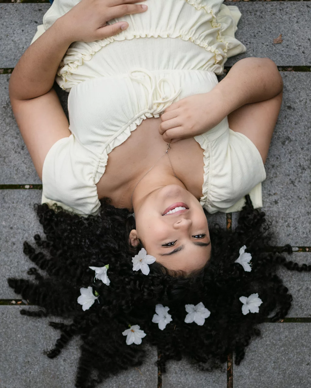 Nolan's senior photos taken at Furman University: A young woman with curly hair lies on her back on a stone surface, smiling up at the camera. White flowers are scattered in her hair, and she wears a light yellow dress.