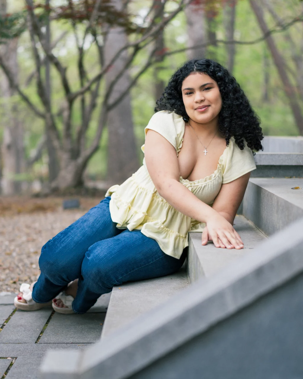 Nolan's senior photos taken at Furman University: A young woman with curly hair, wearing a light yellow blouse and blue jeans, sits on outdoor steps in a wooded area, smiling at the camera.