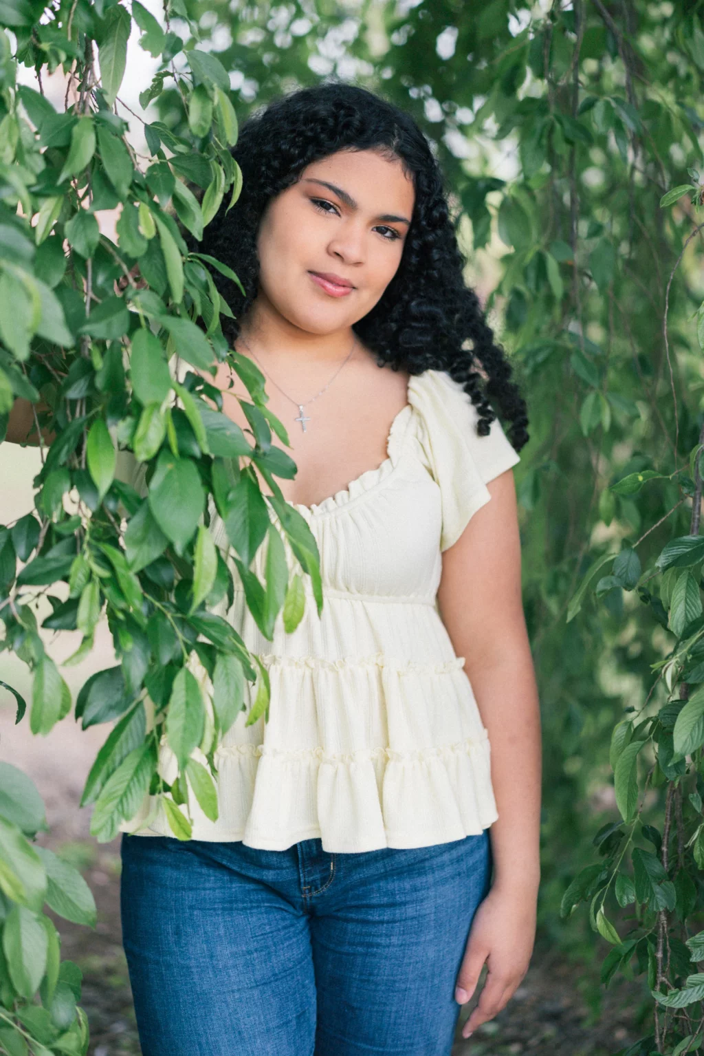 Nolan's senior photos taken at Furman University: A young person with curly hair, wearing a light yellow top and blue jeans, stands among green leafy branches, looking at the camera with a gentle expression.