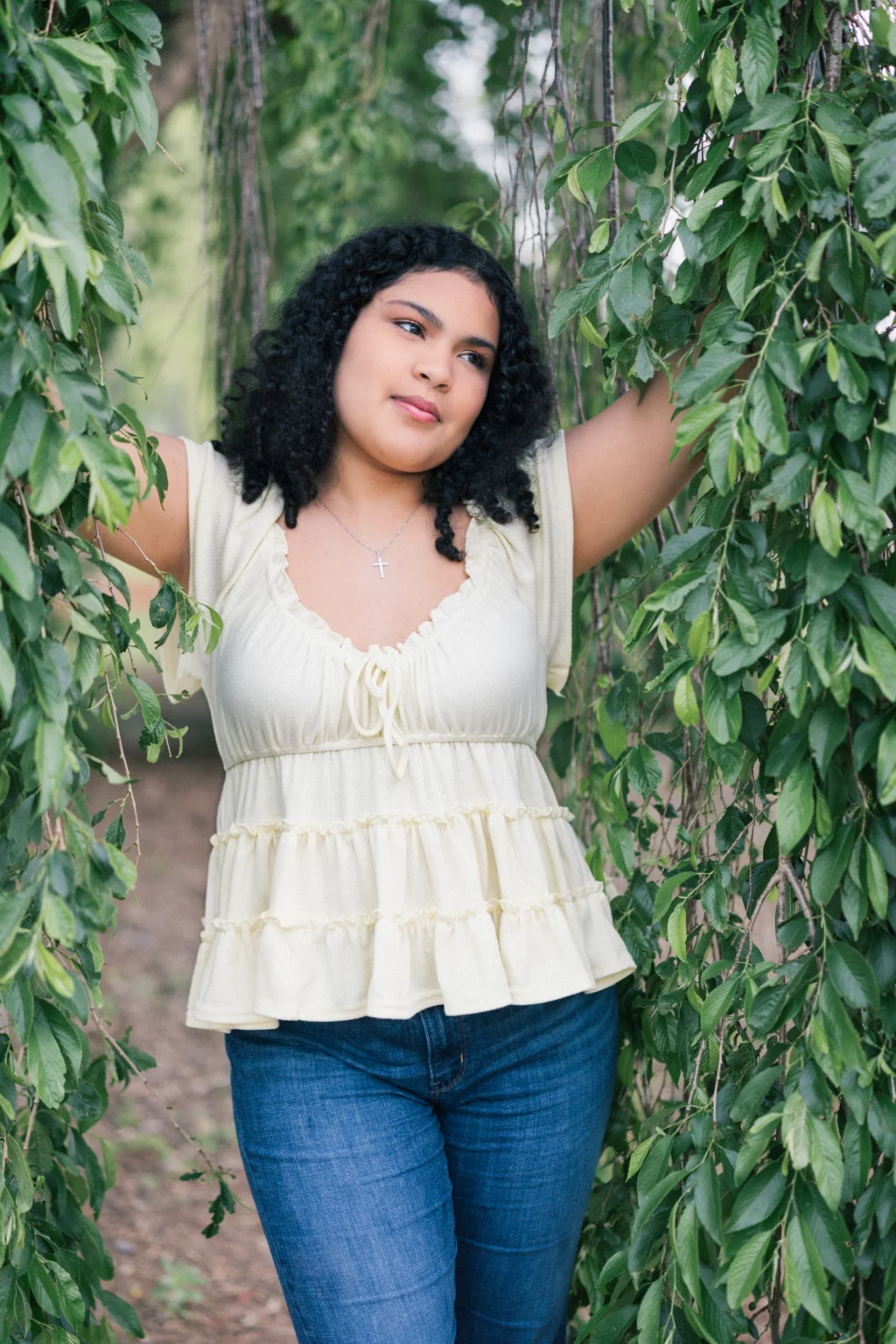 Nolan's senior photos taken at Furman University: A young woman with curly hair stands outdoors, framed by hanging green leaves, wearing a light yellow top and blue jeans, looking thoughtfully to the side.