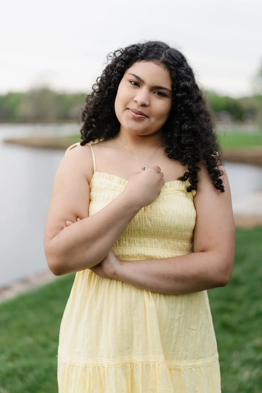Nolan's senior photos taken at Furman University: A young woman with curly dark hair stands outdoors near water, wearing a light yellow sleeveless dress and holding a necklace pendant. Green grass and trees are visible in the background.