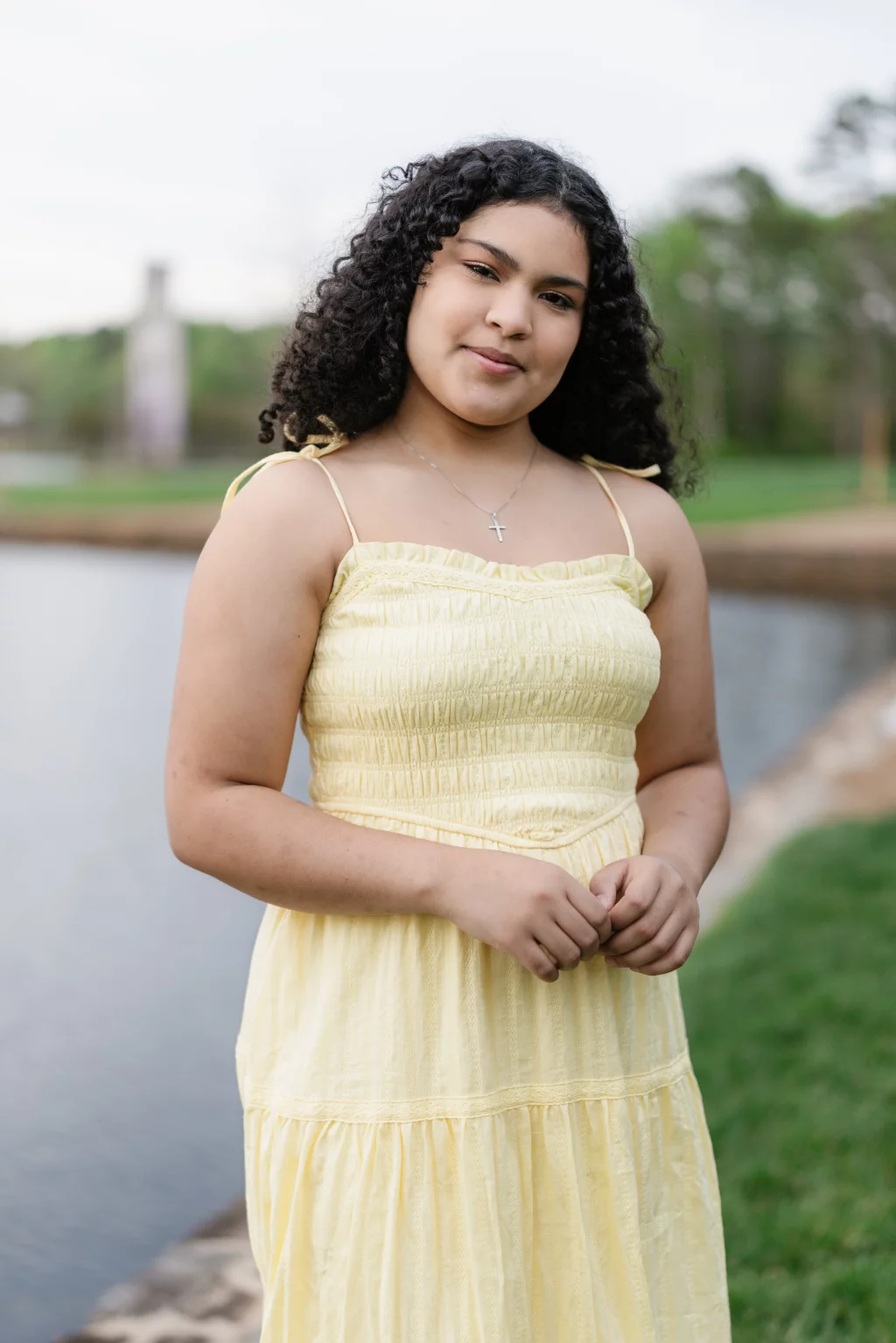 Nolan's senior photos taken at Furman University: Young person with curly hair wearing a yellow sundress and a cross necklace, standing outdoors near a body of water with green grass and trees in the background.