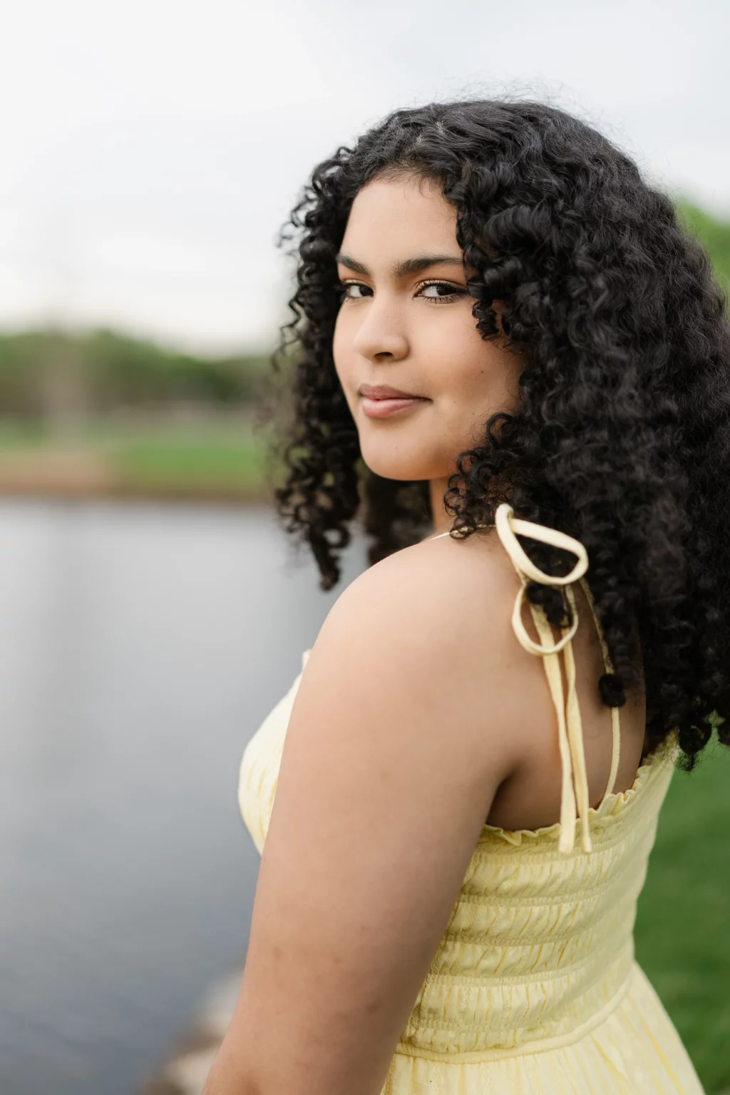 Nolan's senior photos taken at Furman University: A young woman with curly dark hair wearing a yellow sleeveless dress stands outdoors near a body of water, looking over her shoulder at the camera.