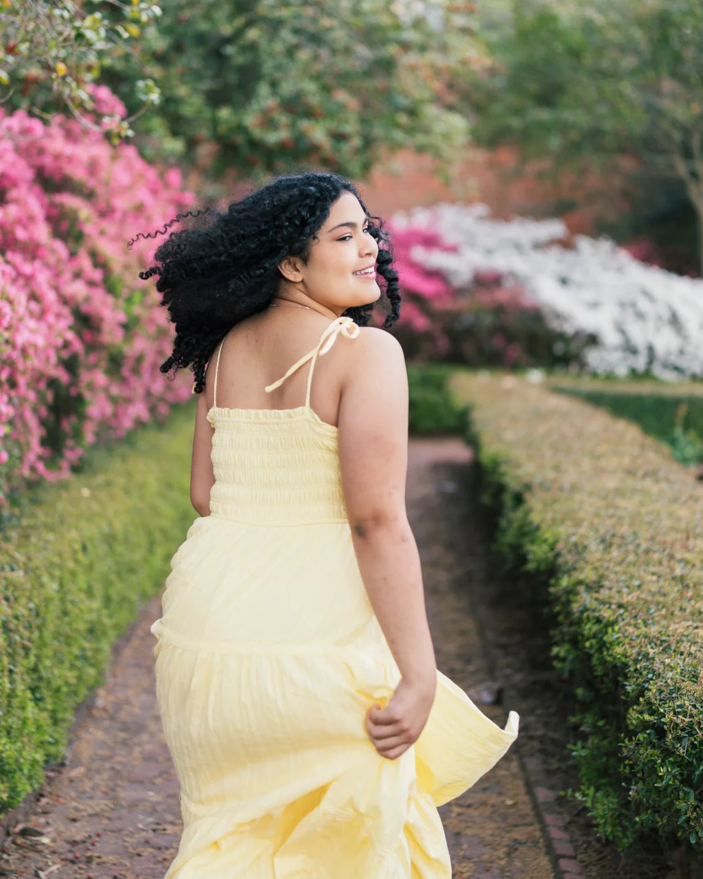 Nolan's senior photos taken at Furman University: Woman with curly hair wearing a yellow dress walks along a garden path lined with green hedges and blooming pink and white flowers, looking back over her shoulder and smiling.