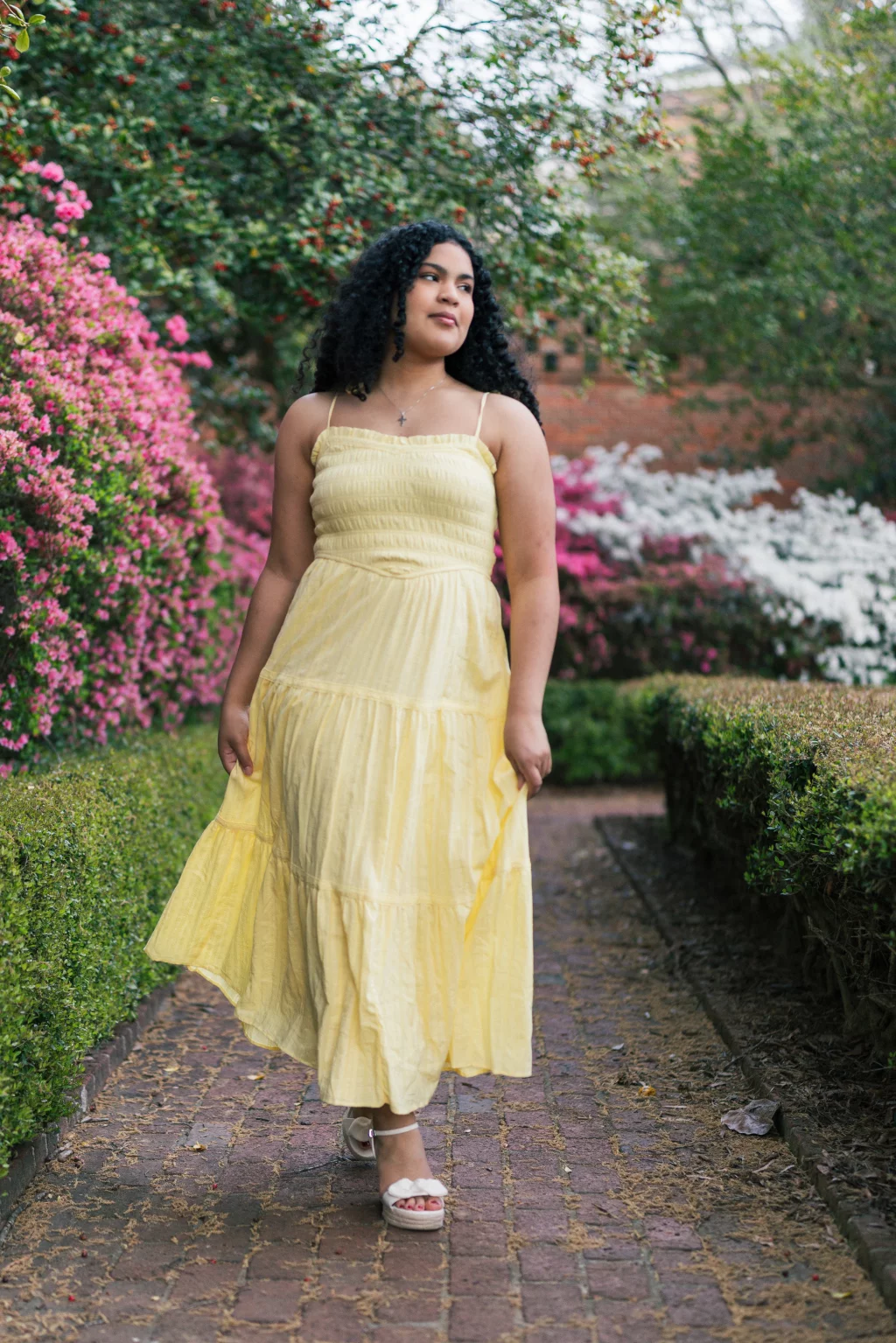 Nolan's senior photos taken at Furman University: A woman in a yellow dress walks along a brick path lined with green hedges and blooming pink and white flowers.