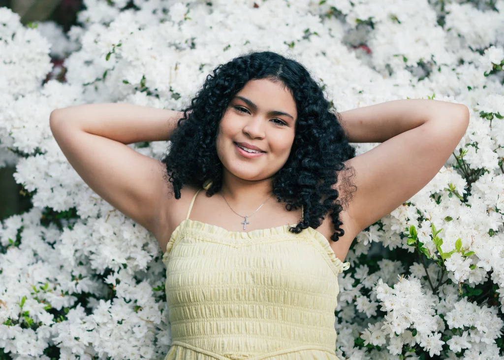 Nolan's senior photos taken at Furman University: Young woman with curly hair wearing a yellow dress and a cross necklace, smiling with arms behind her head, lying against a backdrop of blooming white flowers.