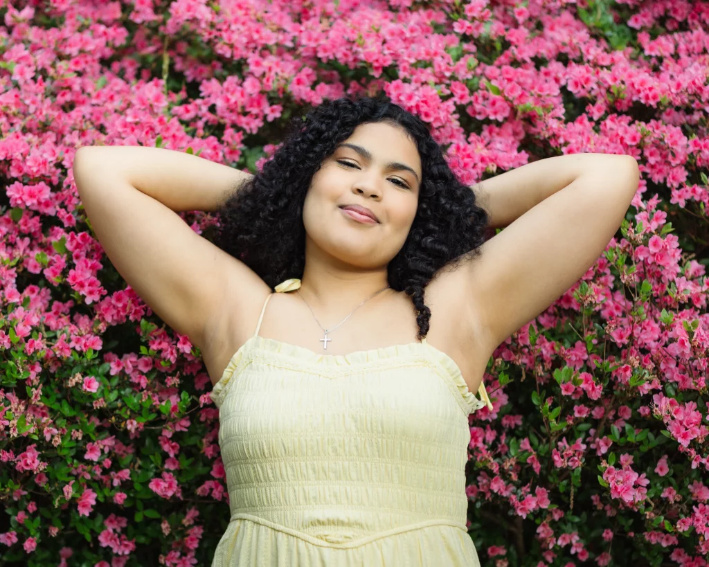 Nolan's senior photos taken at Furman University: Young woman in a light yellow dress standing with arms behind her head, smiling in front of a background of vibrant pink flowers.