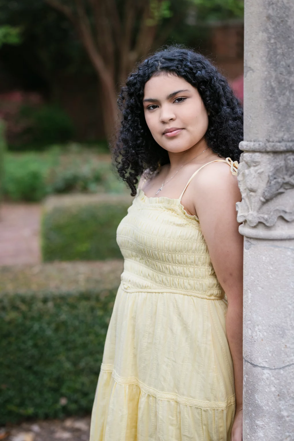 Nolan's senior photos taken at Furman University: A young woman with curly dark hair wearing a light yellow dress stands outdoors, leaning against a stone pillar with greenery in the background.