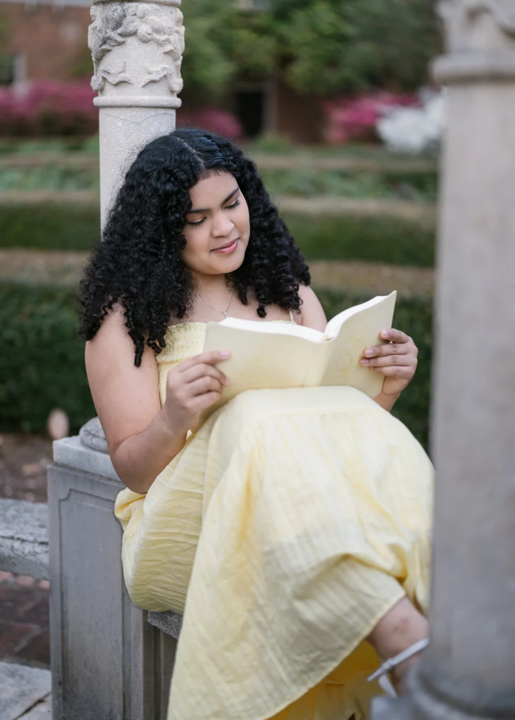 Nolan's senior photos taken at Furman University: A person with curly hair wearing a yellow dress sits outdoors on a stone bench between columns, reading a book and smiling. Manicured hedges and blooming flowers are visible in the background.