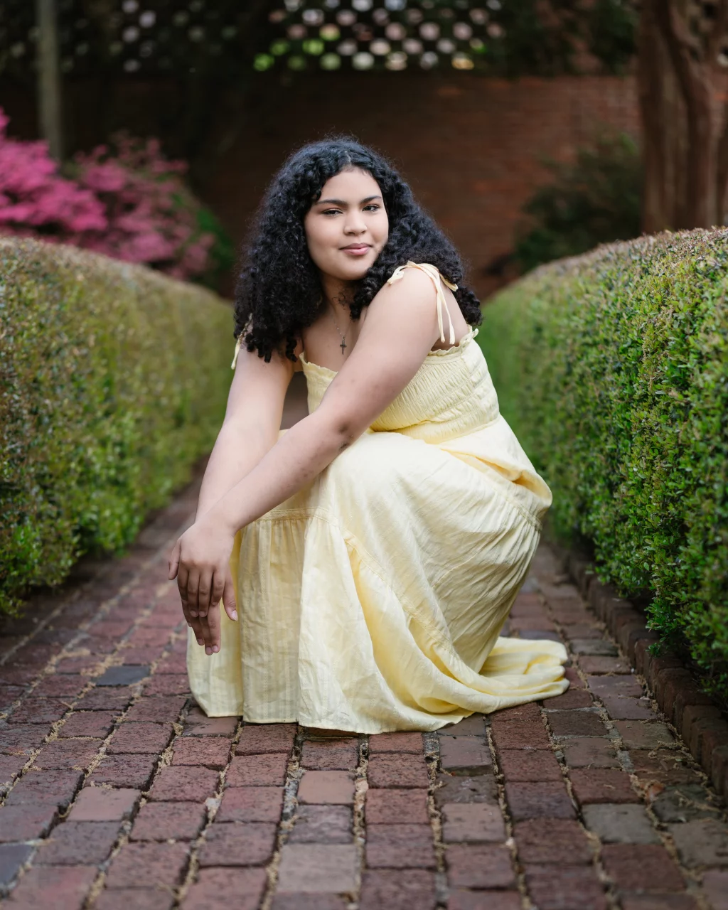 Nolan's senior photos taken at Furman University: A young woman with curly hair wearing a light yellow dress is kneeling on a brick pathway lined with neatly trimmed green hedges, with pink flowering bushes in the background.