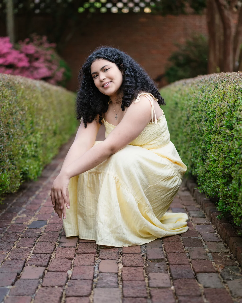 Nolan's senior photos taken at Furman University: Young woman with curly hair wearing a light yellow dress, crouching and smiling on a brick pathway lined with green hedges and pink flowers in the background.