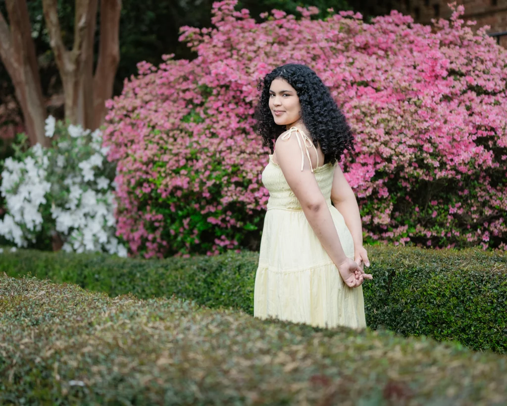 Nolan's senior photos taken at Furman University: A woman with curly hair in a light yellow dress stands in a garden, with pink and white flowering bushes in the background and trimmed green hedges in the foreground.
