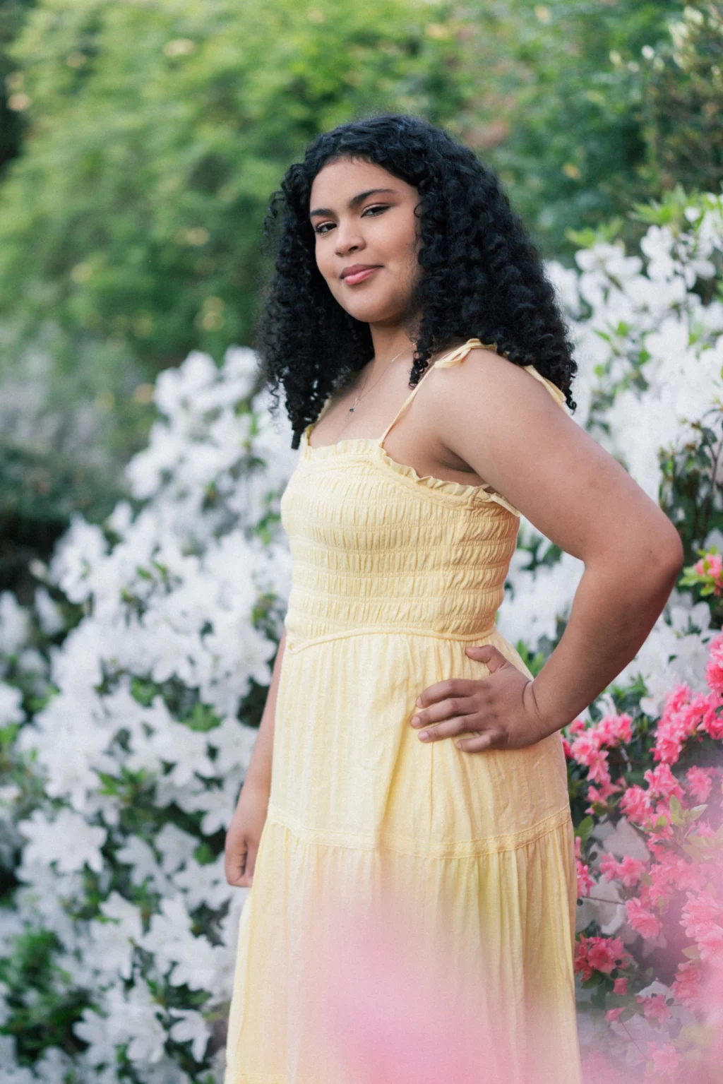 Nolan's senior photos taken at Furman University: Young woman with curly hair wearing a yellow dress stands outdoors in front of blooming white and pink flowers, with greenery in the background.