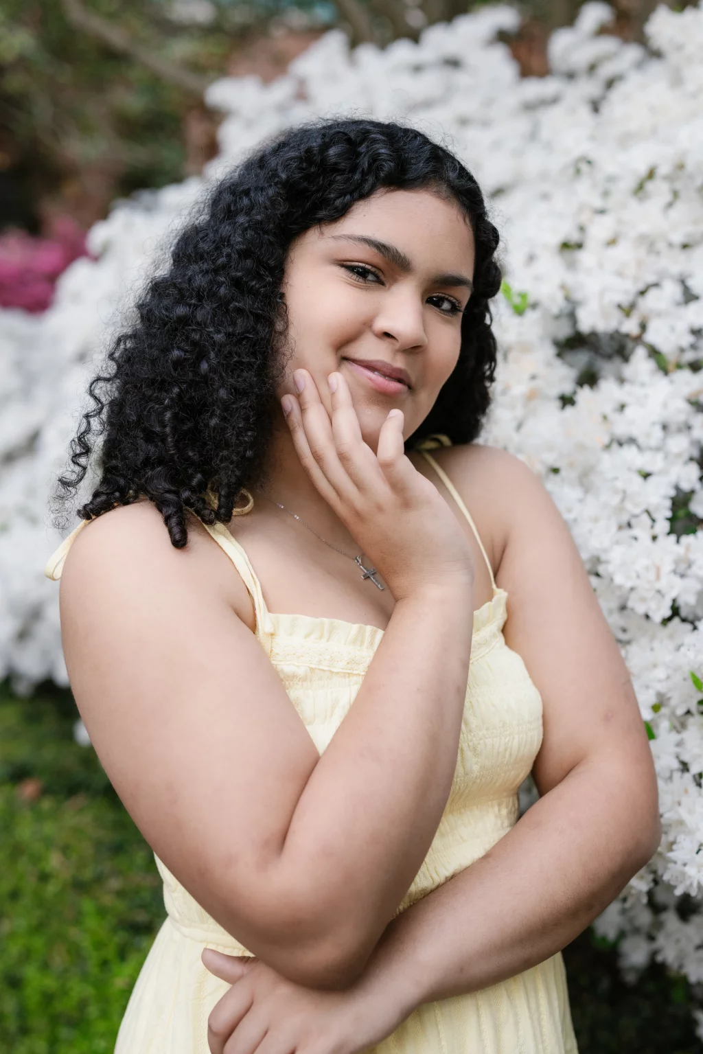 Nolan's senior photos taken at Furman University: Young person with curly hair wearing a light yellow dress, standing in front of white flowering bushes, smiling gently with one hand touching their face.