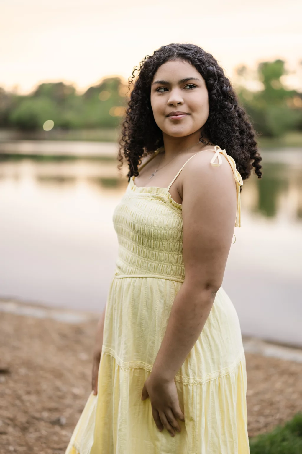 Nolan's senior photos taken at Furman University: Young person with curly hair wearing a light yellow sundress, standing outdoors near a calm lake at sunset.