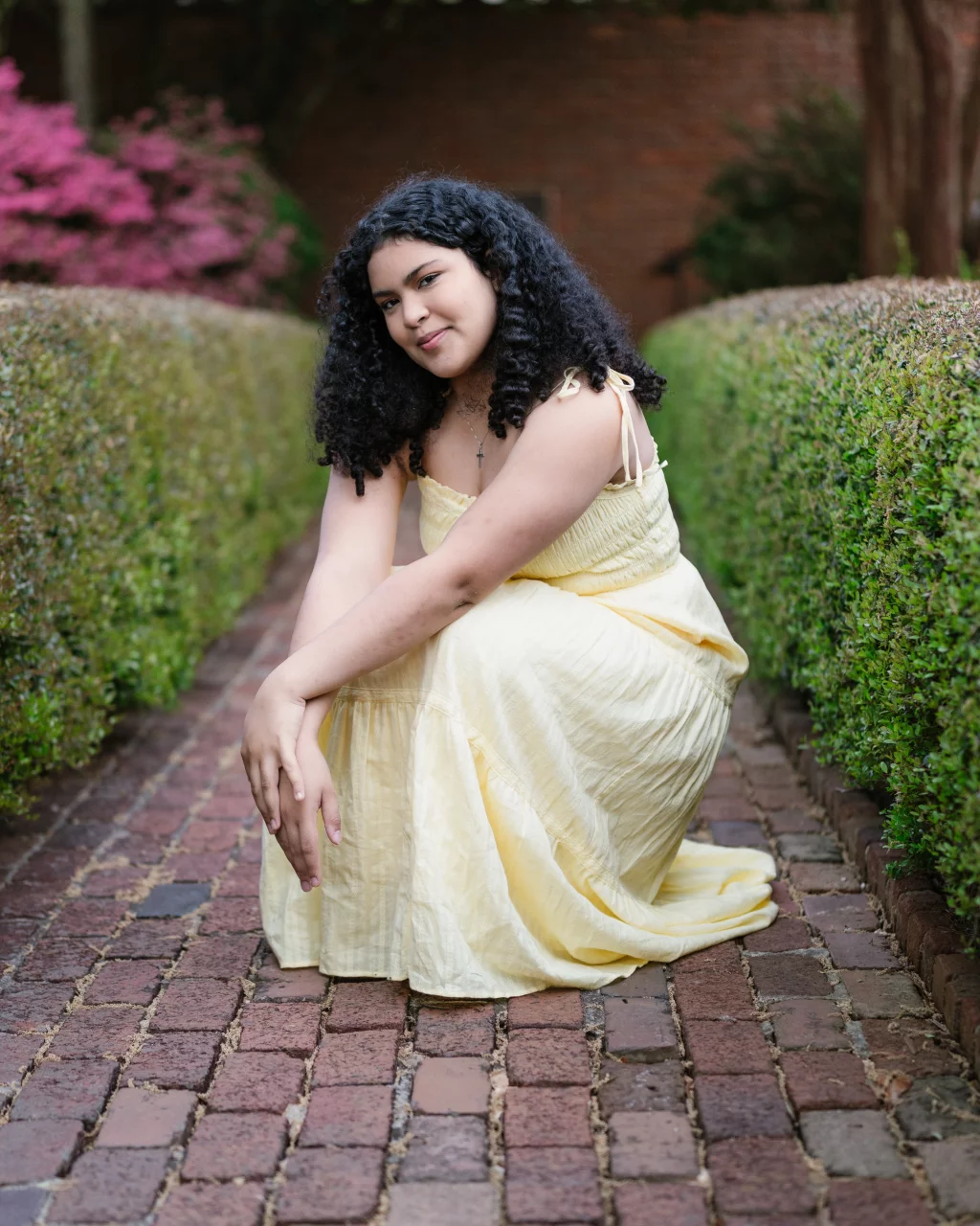 Nolan's senior photos taken at Furman University: A young woman in a light yellow dress kneels on a brick walkway between neatly trimmed green hedges, with pink flowers in the background.