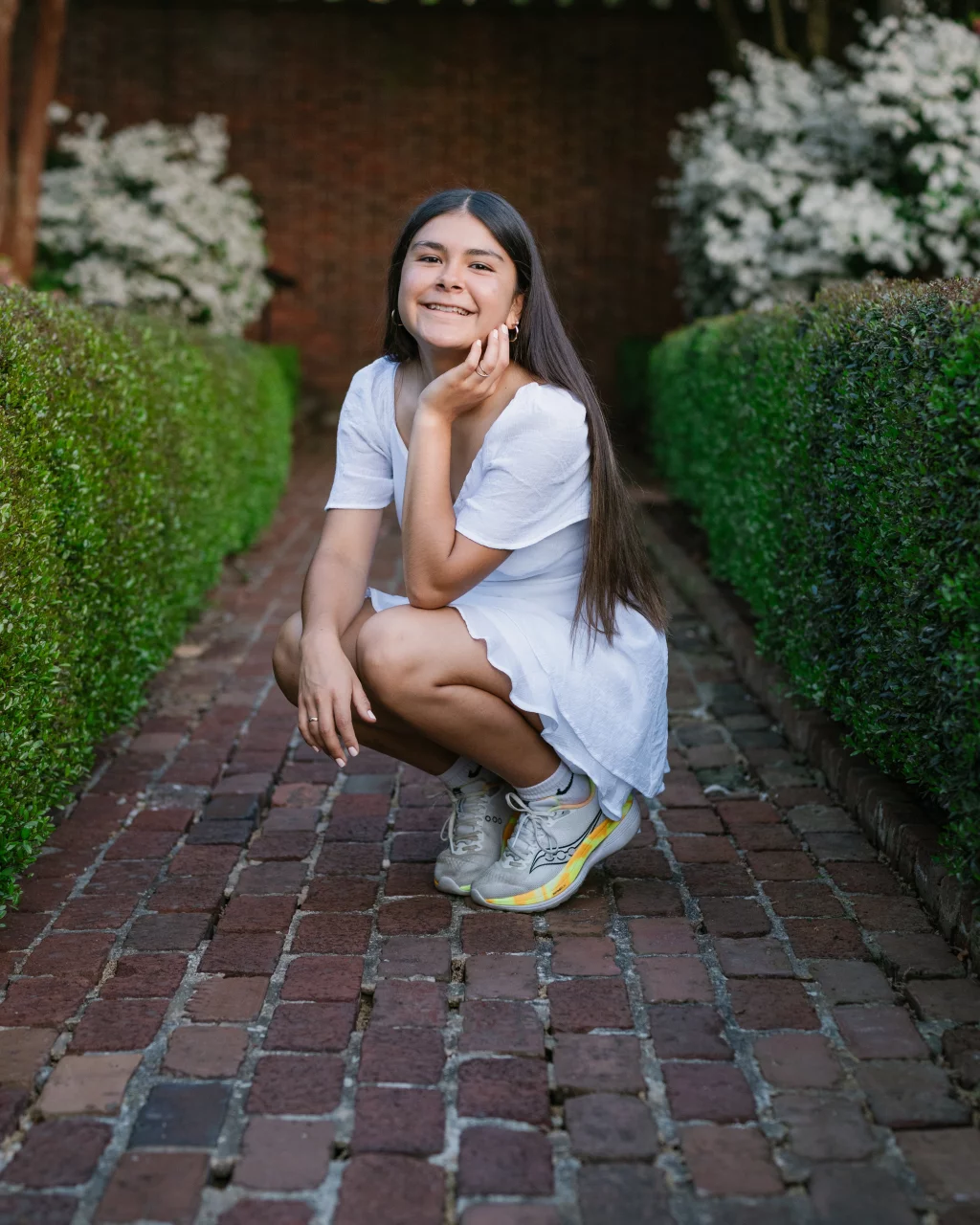 Olivia's senior photos taken at Furman University: Young woman with long dark hair squatting and smiling on a brick pathway, wearing a white dress and sneakers, surrounded by neatly trimmed green hedges and white flowering bushes.