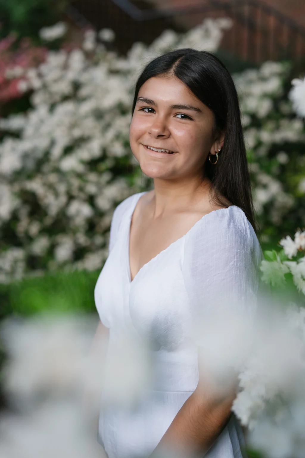 Olivia's senior photos taken at Furman University: A young woman with long dark hair and gold hoop earrings smiles softly, wearing a white dress, standing outdoors in front of blurred white flowers and greenery.