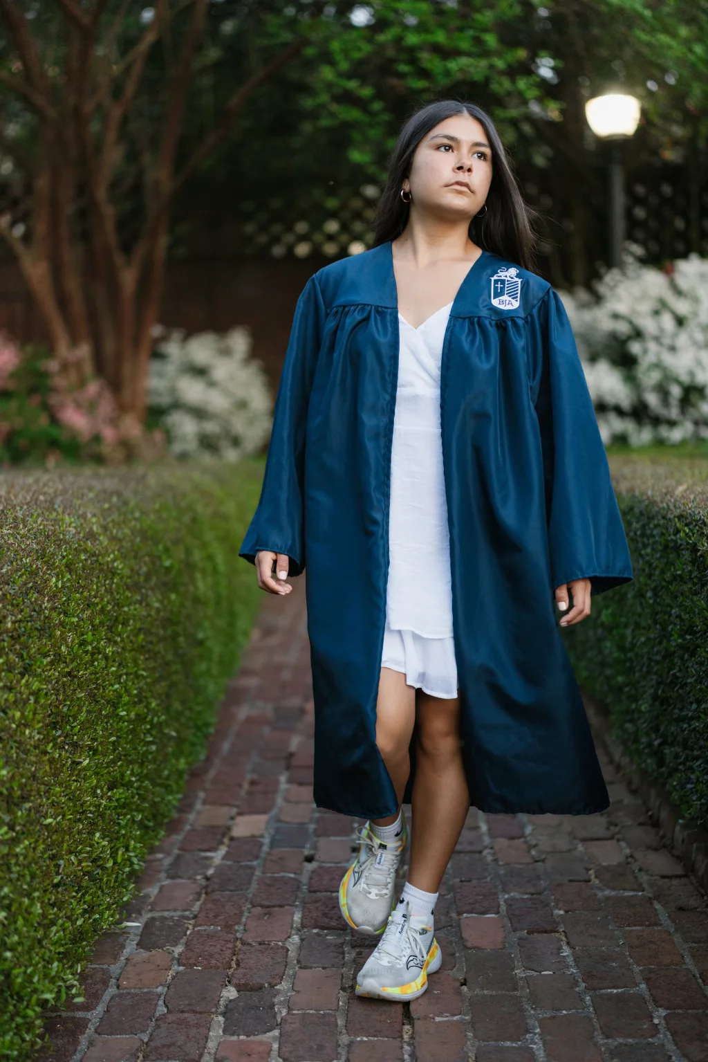 Olivia's senior photos taken at Furman University: A young woman wearing a blue graduation gown over a white dress and sneakers walks on a brick path lined with hedges, with blooming flowers and trees in the background.