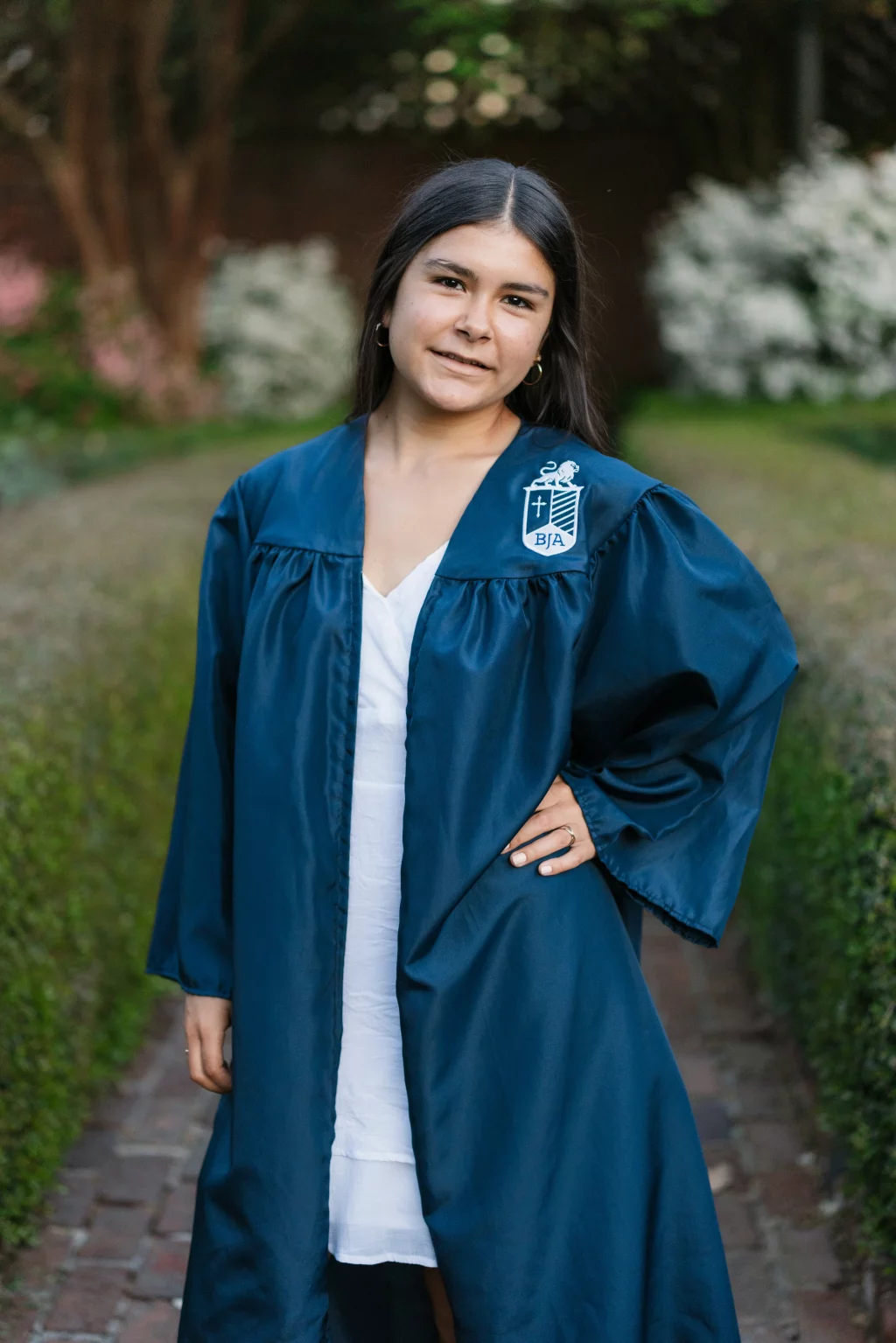 Olivia's senior photos taken at Furman University: A young woman stands outdoors wearing a dark blue graduation gown with a BJA crest, posing with one hand on her hip and a slight smile, surrounded by greenery and flowering bushes.