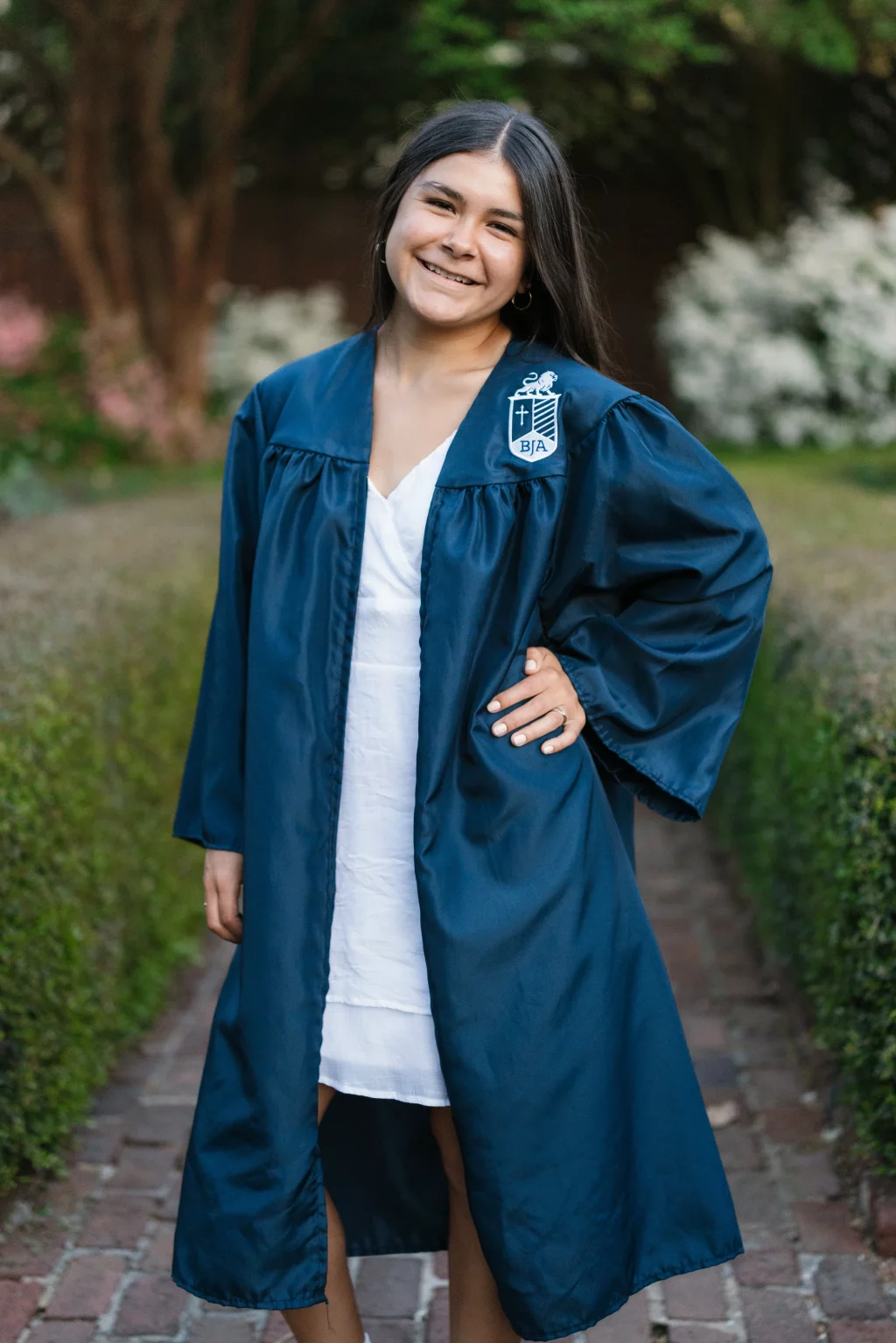 Olivia's senior photos taken at Furman University: Young woman smiling and posing outdoors, wearing a blue graduation gown over a white dress, with one hand on her hip and greenery in the background.