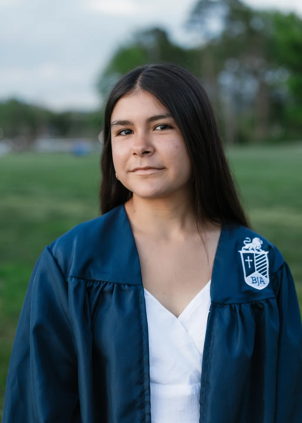 Olivia's senior photos taken at Furman University: Young woman with long dark hair wearing a blue graduation gown with a white emblem, standing outdoors on a grassy field with trees in the background.