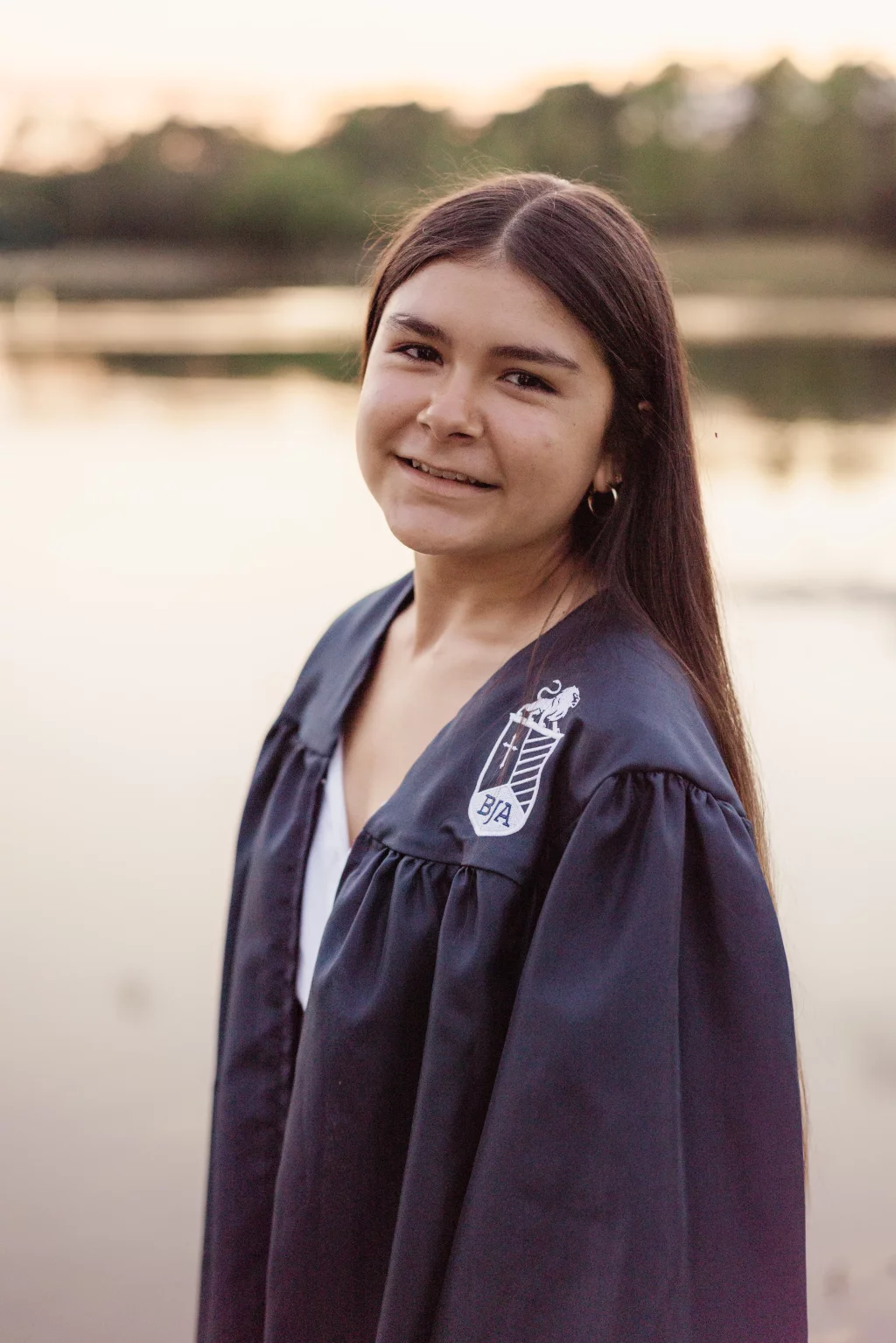 Olivia's senior photos taken at Furman University: Young person in a graduation gown with a crest, standing outdoors by a calm body of water at sunset.