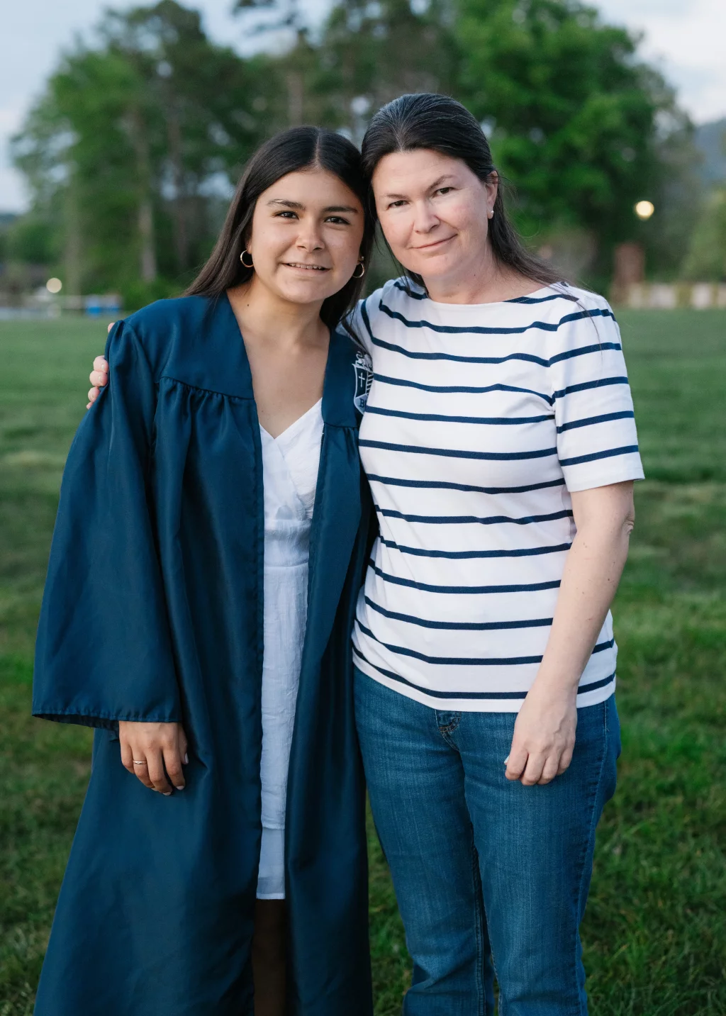 Olivia's senior photos taken at Furman University: Two people standing close together outdoors; one is wearing a dark blue graduation gown and white dress, and the other is in a white and navy striped shirt with jeans. They are smiling and have their arms around each other. Trees and grass are in the background.