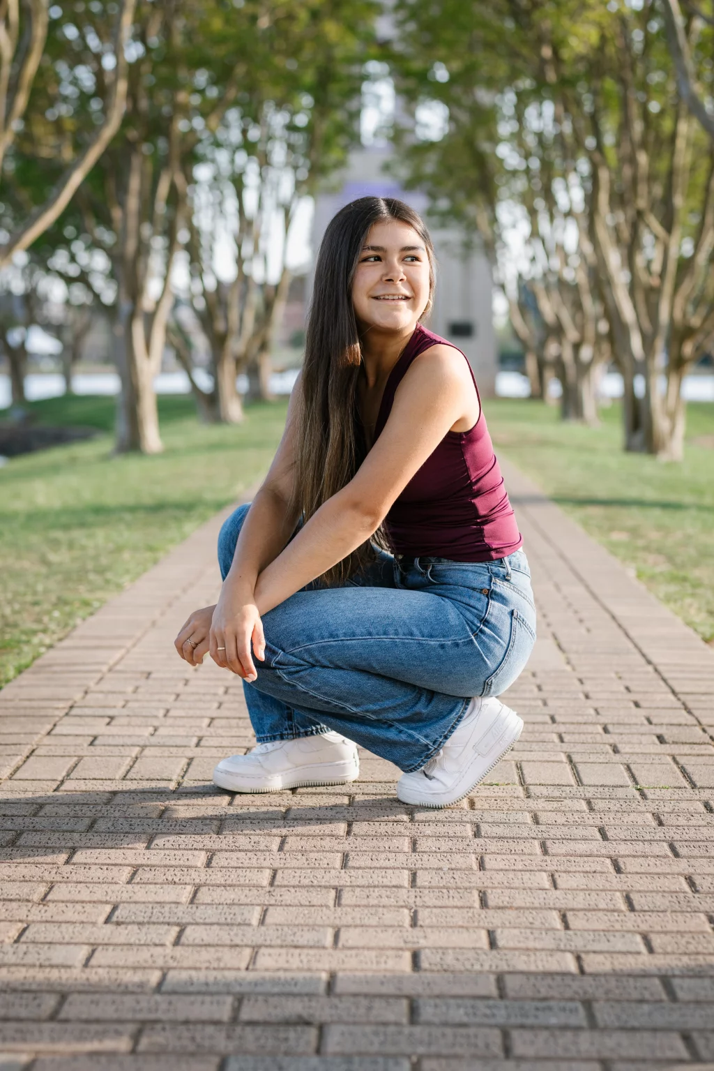 Olivia's senior photos taken at Furman University: Young woman with long hair squatting on a brick pathway in a park, wearing a maroon sleeveless top, blue jeans, and white sneakers, smiling and looking to the side with trees in the background.