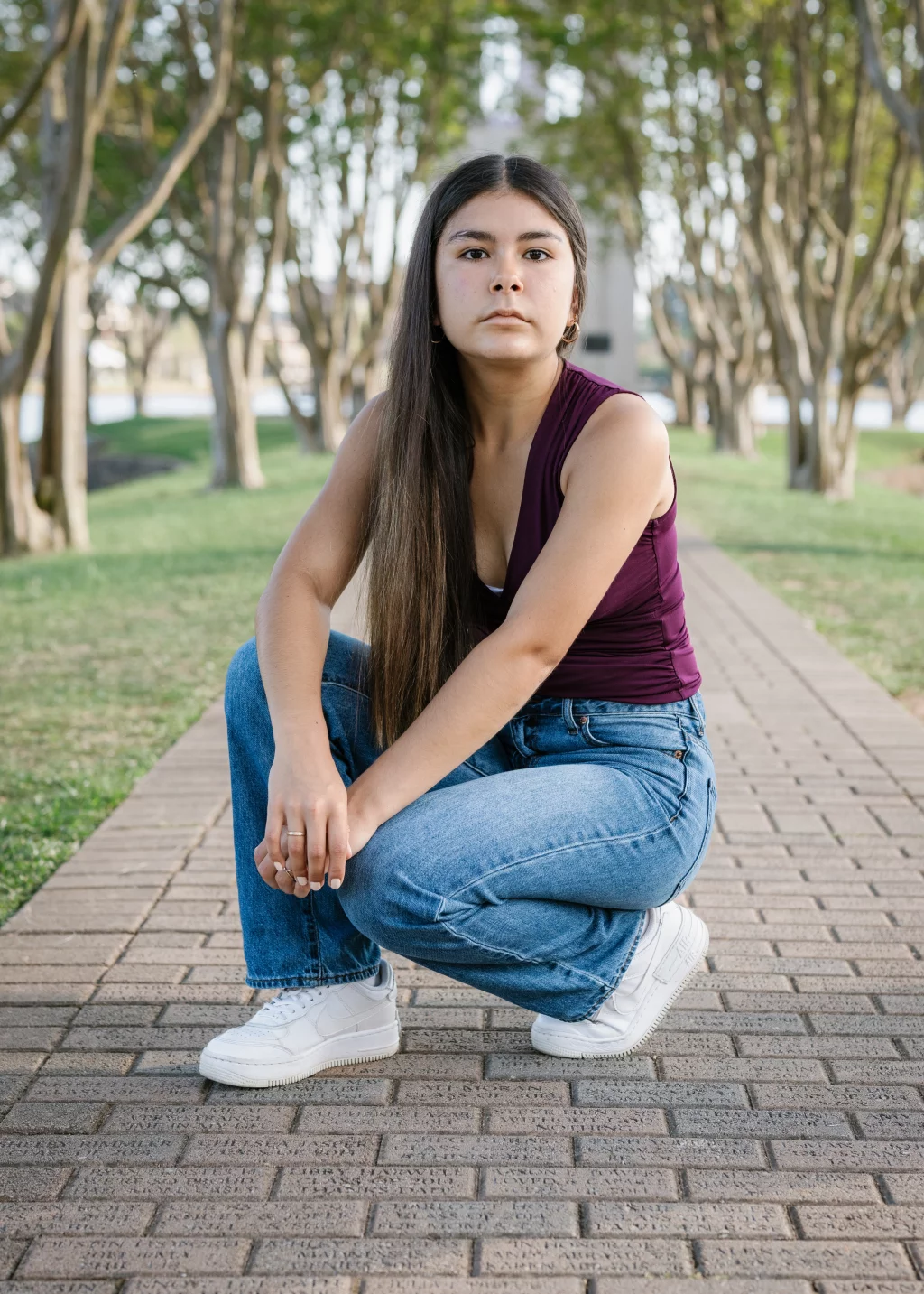 Olivia's senior photos taken at Furman University: A young woman with long dark hair, wearing a sleeveless maroon top, blue jeans, and white sneakers, squats on a brick walkway in a park lined with trees.