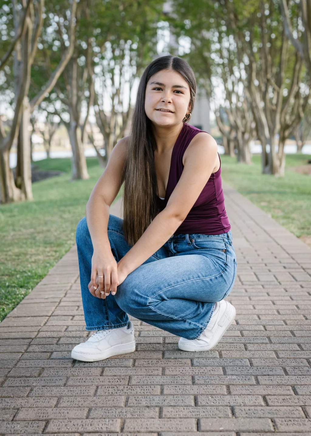 Olivia's senior photos taken at Furman University: A young woman with long straight hair, wearing a sleeveless maroon top, blue jeans, and white sneakers, crouches on a brick pathway lined with trees in a park.