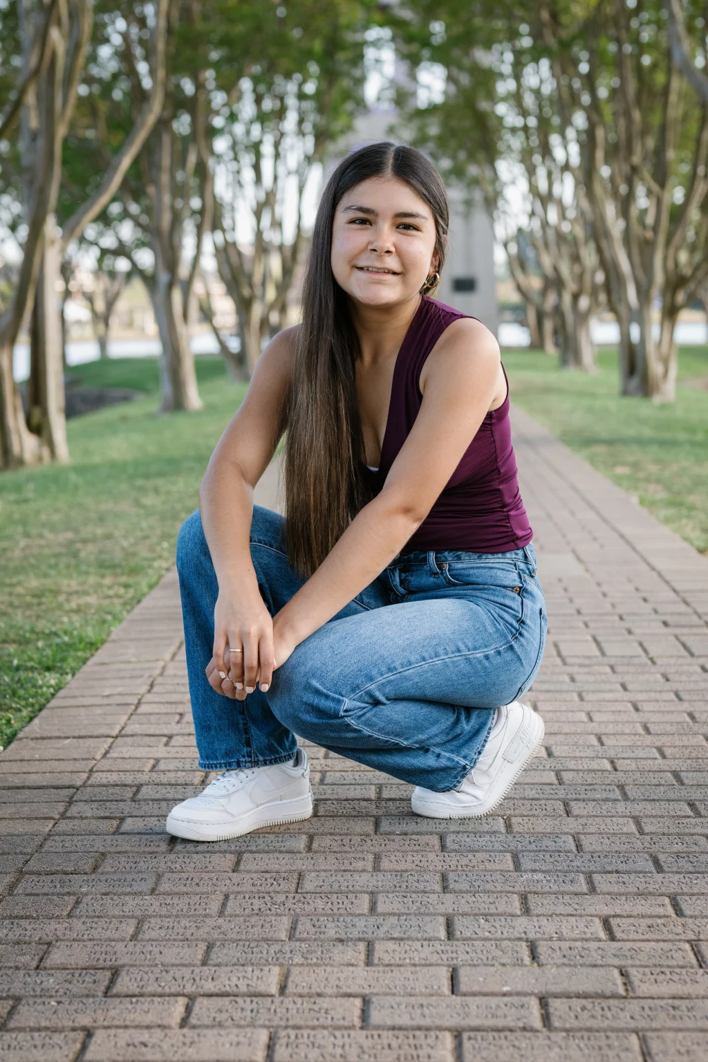 Olivia's senior photos taken at Furman University: Young woman with long dark hair squatting on a brick pathway, wearing a sleeveless maroon top, blue jeans, and white sneakers, with trees and greenery in the background.