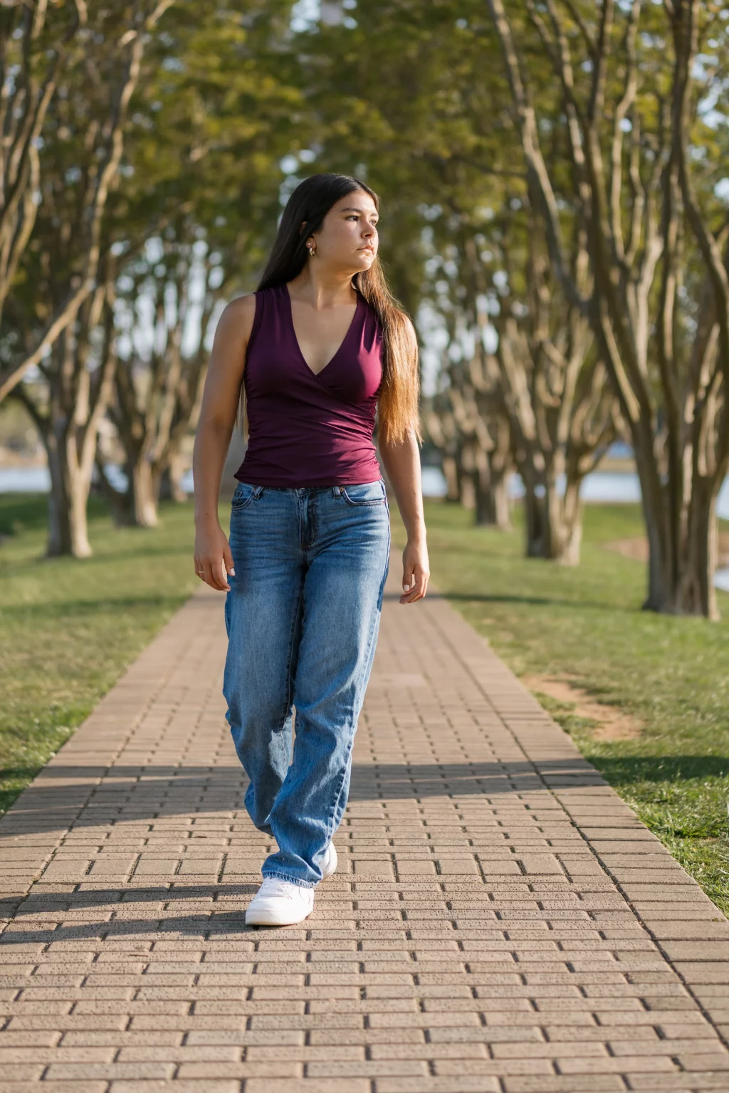 Olivia's senior photos taken at Furman University: A woman with long hair walks on a brick path lined with trees, wearing a sleeveless maroon top, blue jeans, and white shoes.