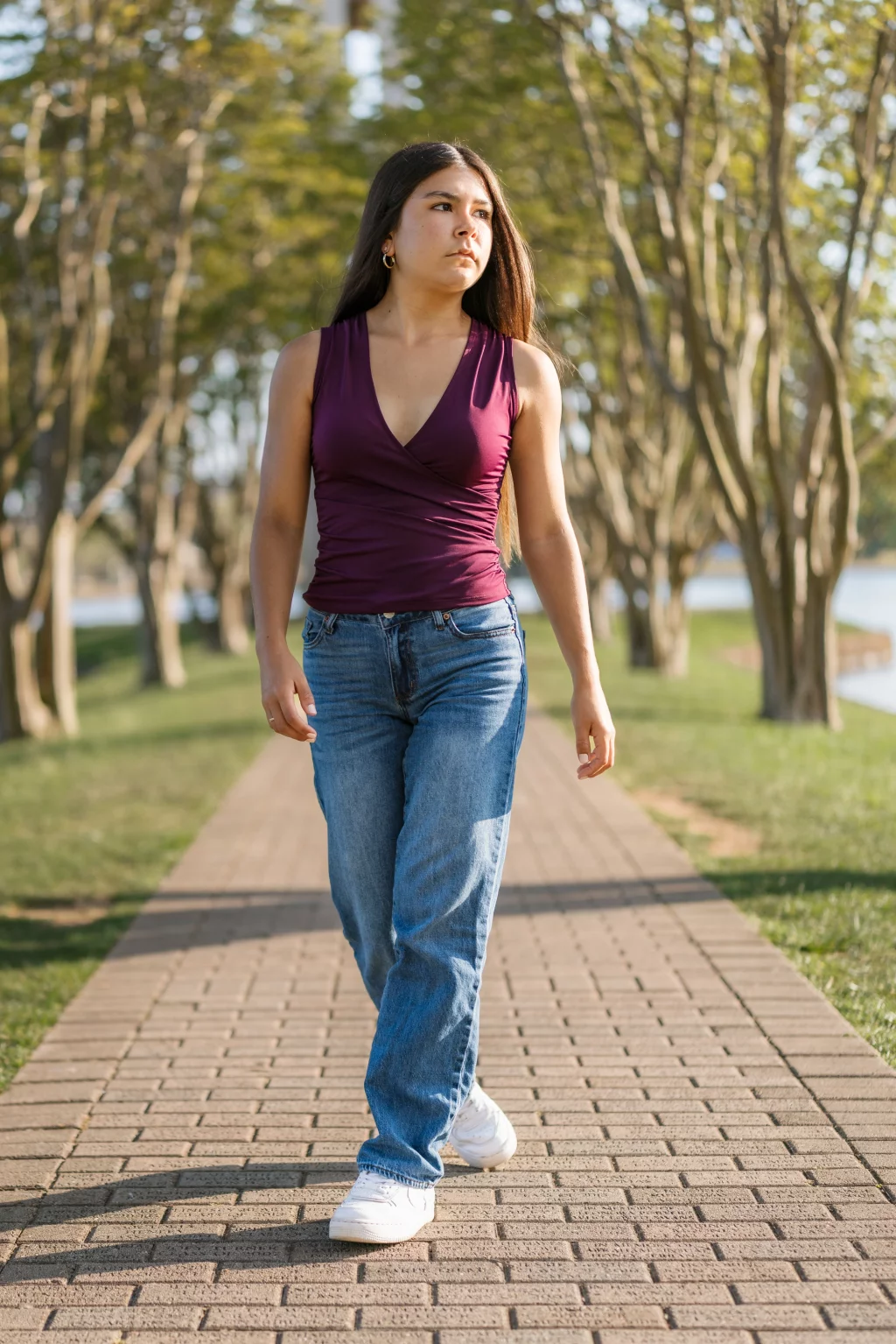 Olivia's senior photos taken at Furman University: A young woman with long dark hair walks on a brick pathway in a park, wearing a sleeveless maroon top, blue jeans, and white sneakers. Trees and green grass line the path, and water is visible in the background.